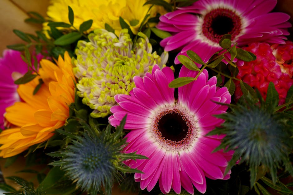 Close-up of a vibrant floral arrangement featuring pink gerbera daisies with dark centers, bright yellow chrysanthemums, green chrysanthemums with pale yellow tips, spiky blue thistles, and lush green