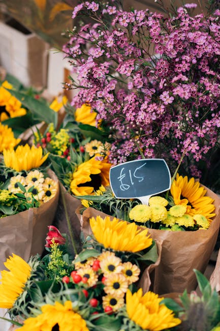 A vibrant display of freshly arranged floral bouquets featuring bright yellow sunflowers, daisy-like blooms, and small red berries, all wrapped in brown kraft paper. The flowers are placed outdoors on