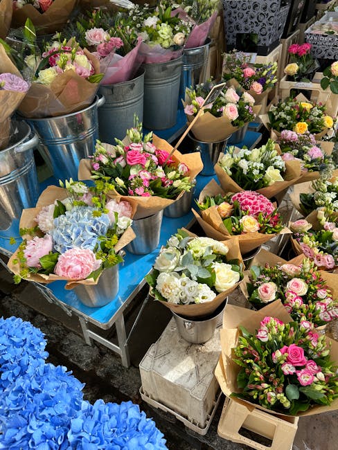A variety of freshly arranged floral bouquets displayed on tables and in metal buckets, featuring roses, hydrangeas, and other seasonal flowers in shades of pink, white, purple, and blue. The bouquets