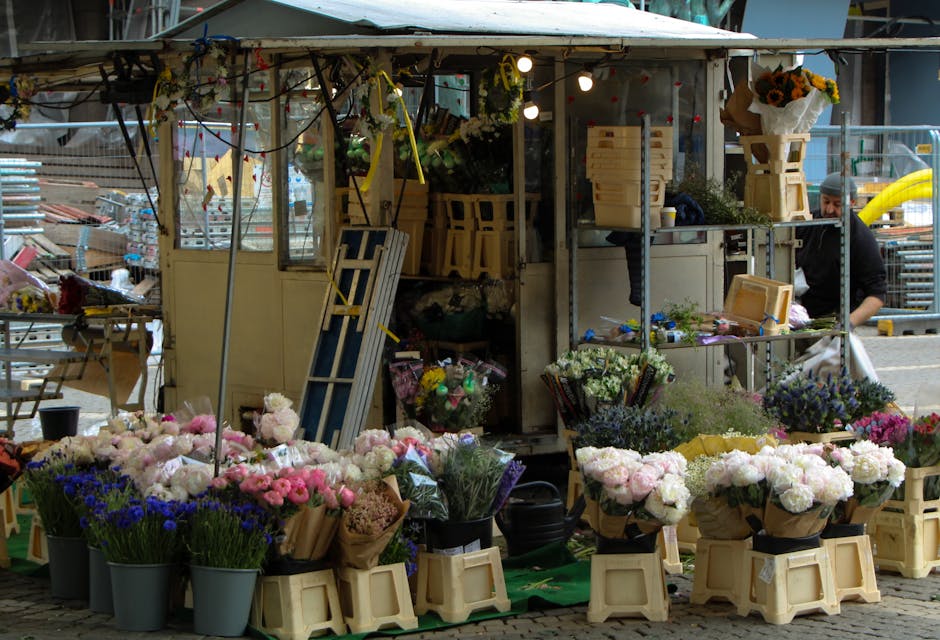 A vibrant flower stall set up outdoors, featuring an array of fresh floral arrangements including pink, purple, and white blooms, all displayed in buckets and wrapped in brown paper. The arrangement i