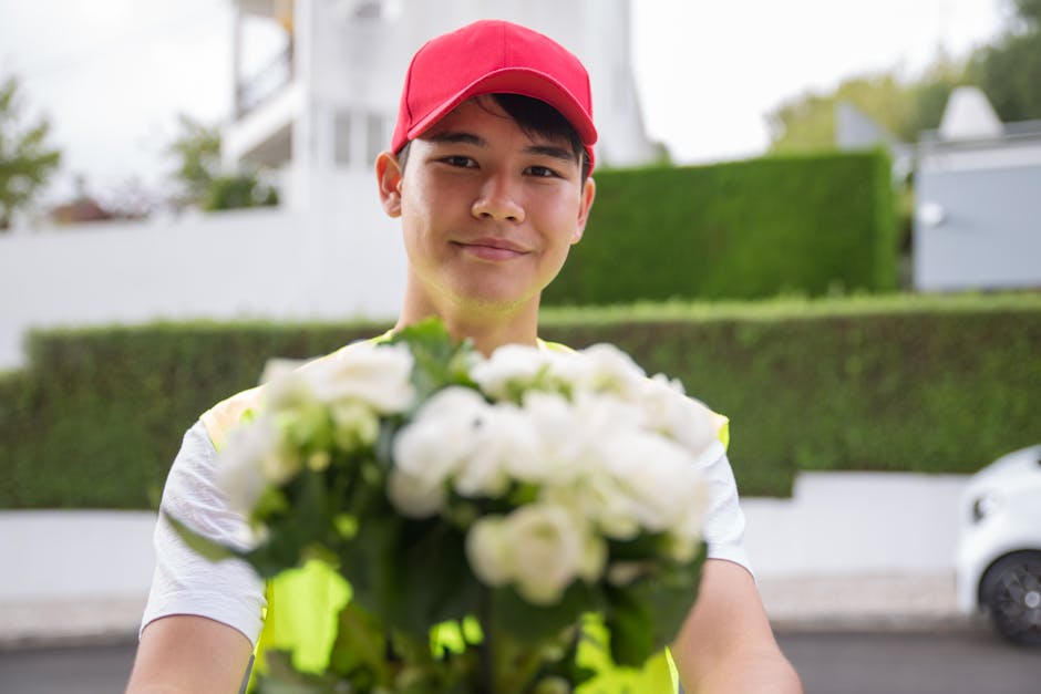 A delivery person wearing a red cap and a high-visibility vest holds a vibrant bouquet of fresh white roses and green foliage, extending it towards the camera. The bouquet is wrapped in clear cellopha