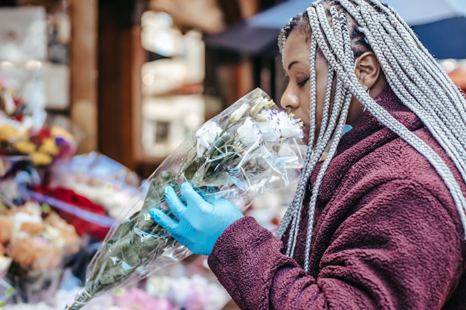A woman with intricate silver braids and wearing a maroon fleece jacket is holding a bouquet of white roses wrapped in clear cellophane at an outdoor flower market. She is wearing a light blue glove o