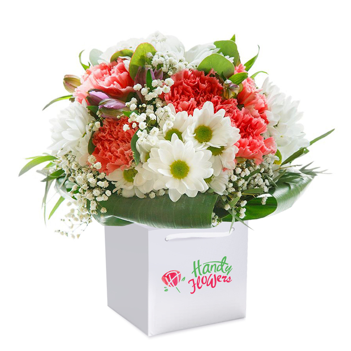 Presented upright in a crisp white square gift bag, this eye-catching bouquet embodies both freshness and artistry, designed with Morden residents in mind. At its core, effervescent coral and peach carnations blossom with fullness and depth, their ruffled petals forming waves of texture reminiscent of the tranquil gardens near the Wandle Trail. Alongside these, brilliant white daisies with vivid lime-green centres make a striking contrast, reminiscent of the cheerful, sunlit scenes at Morden Hall Park. Scattered delicately among the central blooms, clusters of airy white baby's breath provide a soft, cloud-like frame, while the addition of a few closed, purple-hued buds on one side adds dimension and a sense of unfolding potential. The supporting cast of greenery-broad glossy leaves and smaller forest-green fronds-encircles the arrangement, forming a natural, elegant wrap that enhances the sense of abundance. The overall shape is a harmonious dome, carefully structured to express both generosity and precise florist skill. The immaculate white gift bag, outfitted with sturdy white ribbon handles for easy lifting, emphasizes a modern approach to gifting without distracting from the bouquet's vibrant colours. Set against a flawless white background, the entire ensemble radiates with a sense of occasion-perfect for birthdays, anniversaries, or a spontaneous gesture to brighten any home from Martin Way to Morden South. Every petal, stem, and leaf is selected to reflect quality, care, and the distinctive charm of floral gifts available locally throughout Morden.